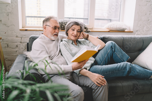 Elderly couple reading book together while sitting on sofa at living room