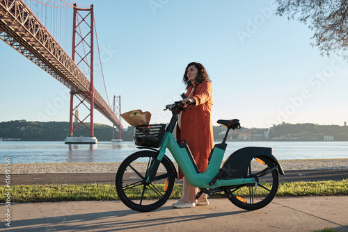 a Pregnant woman in orange coat on electric bicycle on the promenade in Lisbon near the bridge on April 25