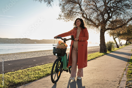Fotografie Pregnant woman in orange coat on electric bicycle on the promenade at sunset