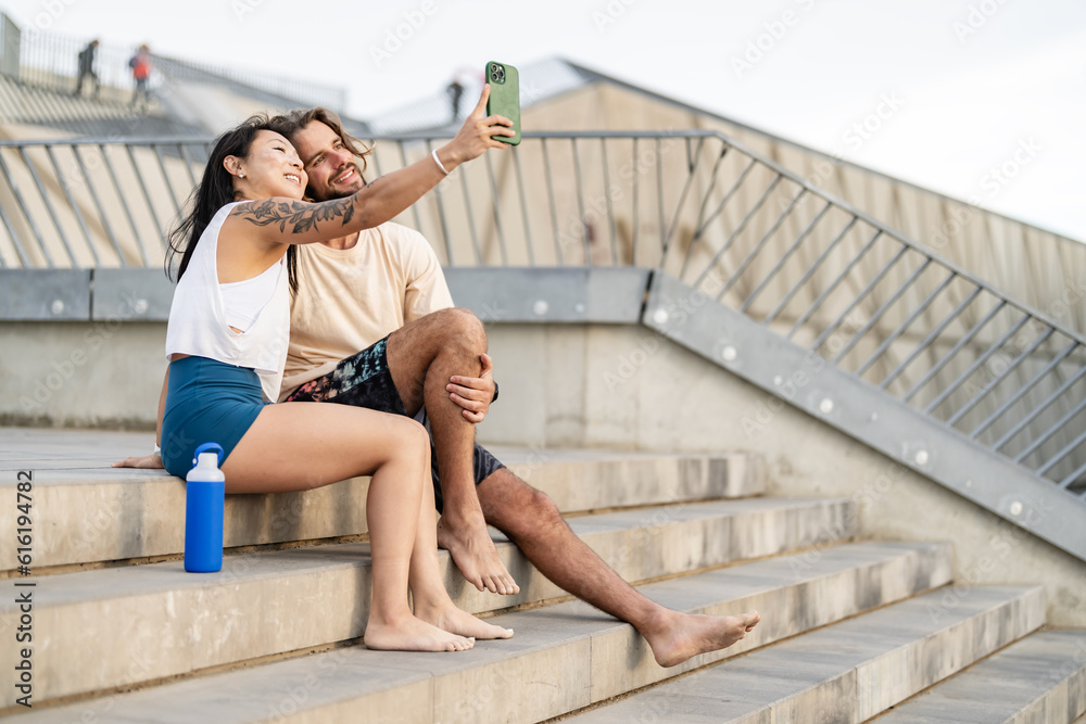 Young diverse urban couple sitting on stairs outdoors in the city, looking at a smartphone, communicating, having fun