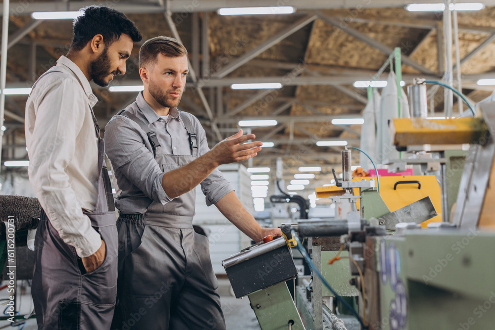 A male inspector or operator of a workshop for the production of aluminum and plastic wreaths trains an intern. International team of men working together near a machine in a factory.