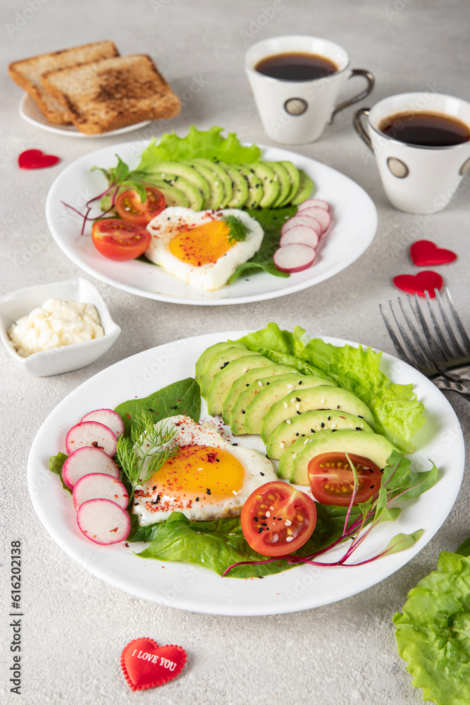 Romantic couple breakfast - two portions heart-shaped fried eggs with avocado, spinach, cherry tomato and radish on plates and two cups of coffee
