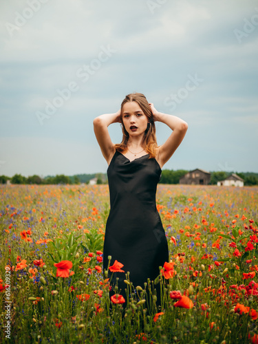 Beautiful young girl in a black evening dress posing against a poppy field on a cloudy summer day. Portrait of a female model outdoors. Rainy weather. Gray clouds. Vertical shot.