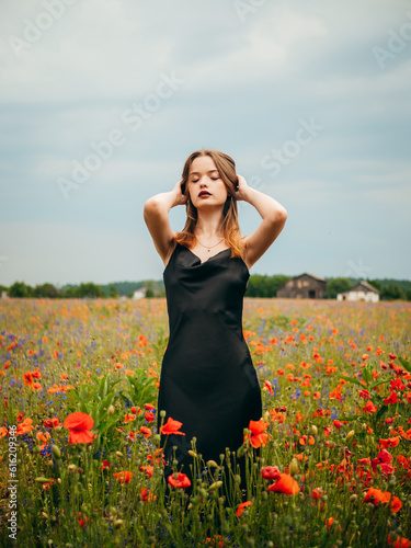 Beautiful young girl in a black evening dress posing against a poppy field on a cloudy summer day. Portrait of a female model outdoors. Rainy weather. Gray clouds. Vertical shot.