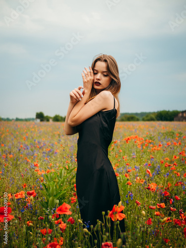 Beautiful young girl in a black evening dress posing against a poppy field on a cloudy summer day. Portrait of a female model outdoors. Rainy weather. Gray clouds. Vertical shot.