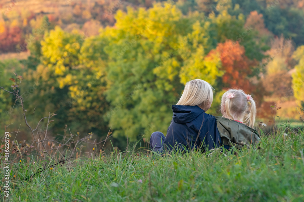 Two siblings sit hugging on grass on autumn landscape background. Carefree childhood. Back view Two siblings sit hugging on grass on autumn landscape background. Carefree childhood. Back view