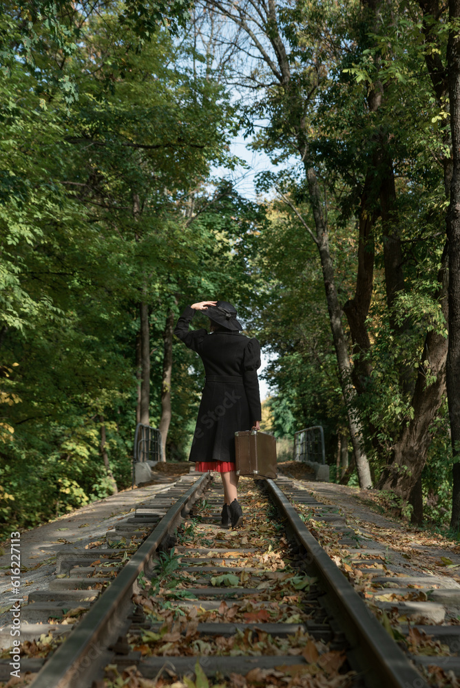 Back view on lady in black coat and hat of 40s style with valise in hands walks along rails in forest. Back view