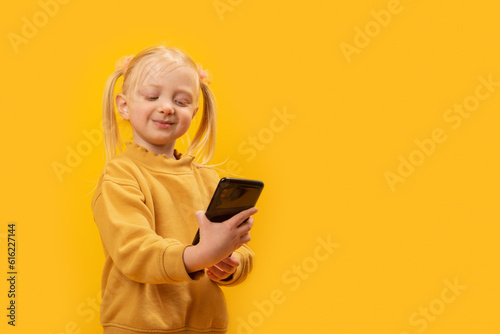 Cute fair-haired kid with two ponytails in yellow jacket looks at phone and smiles. Preschooler girl takes selfies.