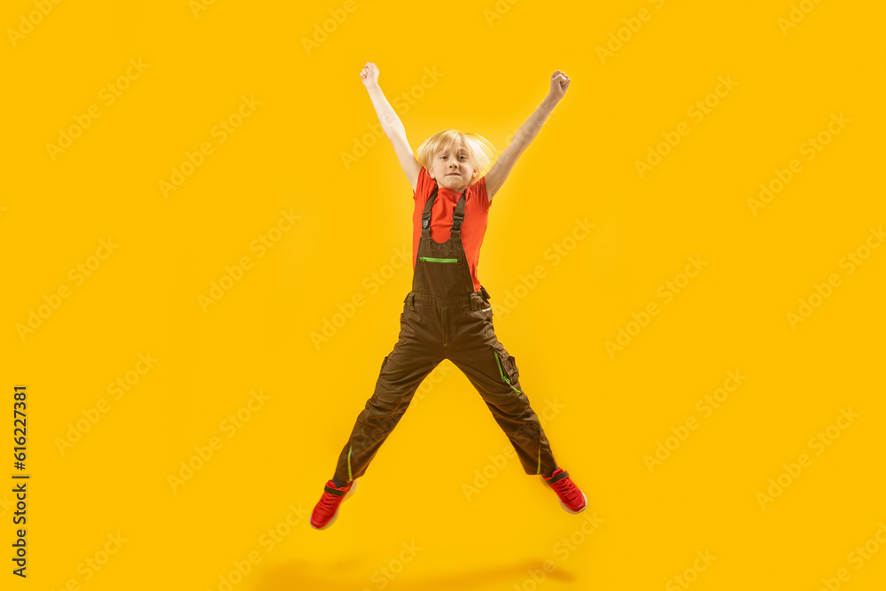 Blond boy jumps high with his hands up. Portrait of schoolboy in overalls bouncing on yellow background. Copy space.