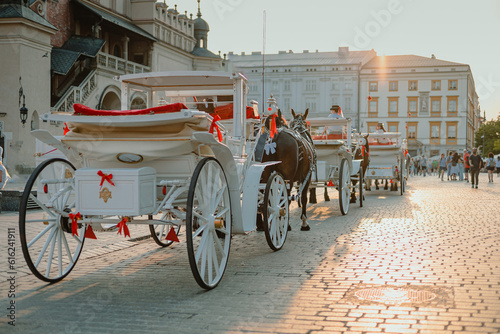 Photography White fancy wooden horse drawn carriages on the Krakow main square or Rynek Glowny on a romantic sunny afternoon