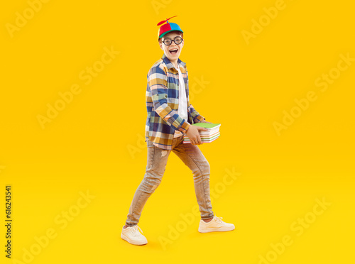 Funny school child carrying books. Full body shot of happy little boy in casual clothes, round glasses and propeller cap smiling, holding pile of books and walking on yellow studio background