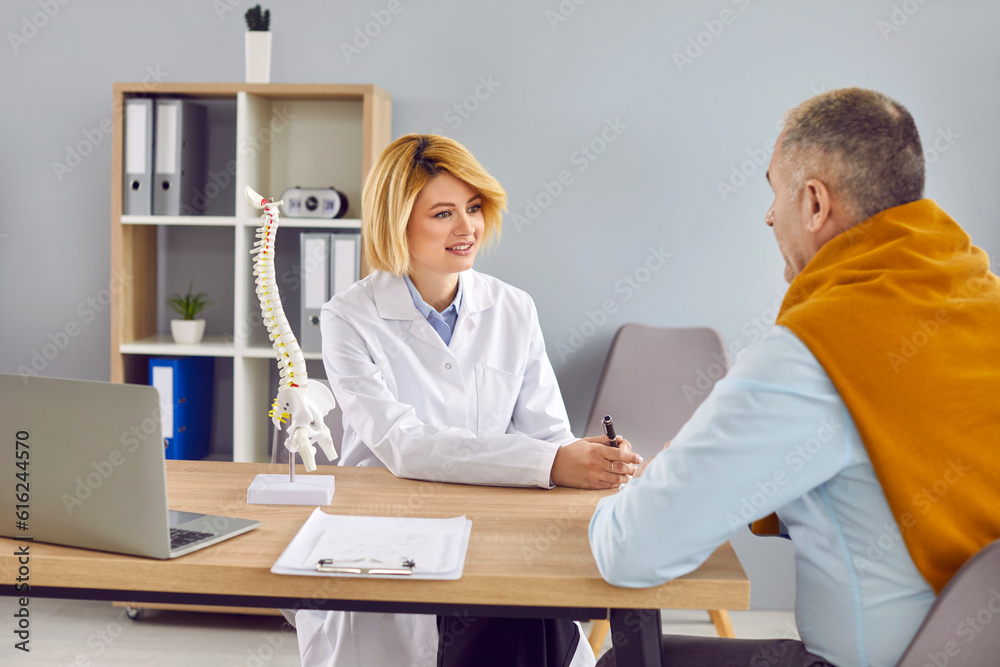 Obraz premium Young woman doctor orthopedic surgeon in medical white coat, with laptop and spine model on desk, talking to senior man patient, explaining something, telling about skeleton characteristics