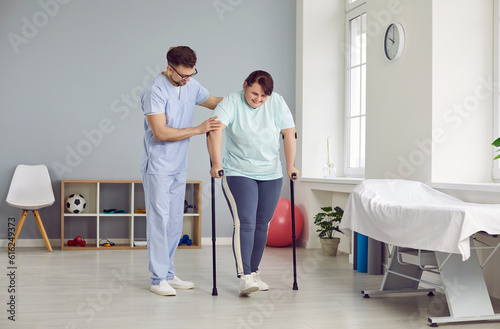 Fényképezés A young cheerful friendly nurse helping fat plus size female patient to walk with her crutches in medical clinic