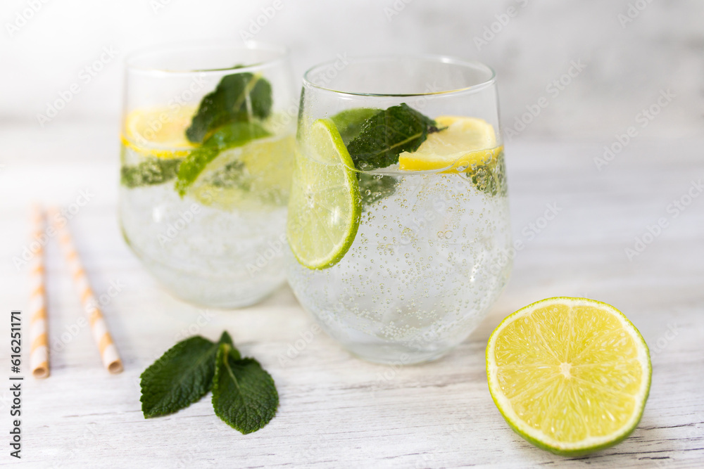 Fototapeta premium Cold refreshing lemonade drink or cocktail made of sparkling water, citrus and mint. Served drinking glass with ice cubes. On white wooden backgrond with copy space. 