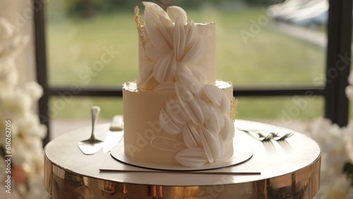 Elegant luxury two tiered white wedding cake decorated edible flowers, dry rustic branches and icing on stand with golden decor at reception restaurant, ceremony arch on background. Slow motion