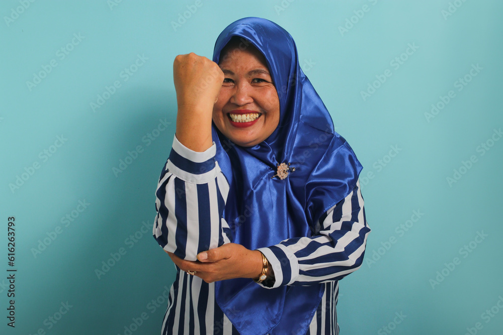A smiling middle-aged Asian woman, in a blue hijab and striped shirt, makes a strong gesture by lifting her arms and flexing her muscles towards the camera, standing against a blue background A smiling middle-aged Asian woman, in a blue hijab and striped shirt, makes a strong gesture by lifting her arms and flexing her muscles towards the camera, standing against a blue background