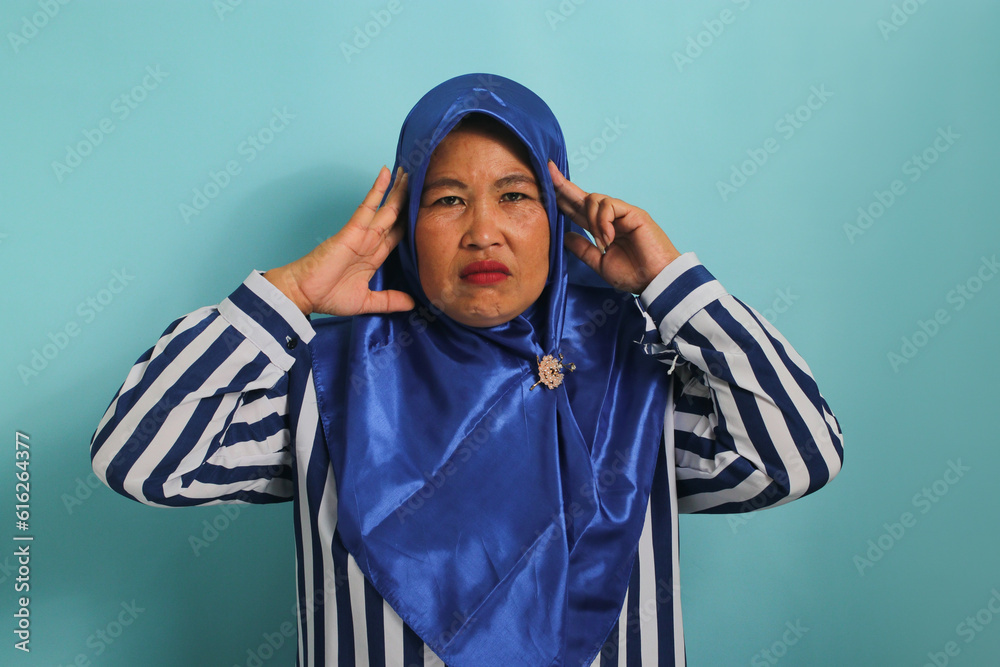 A troubled middle-aged Asian woman, wearing a blue hijab and a striped shirt, grabs her head and stares alarmed at the camera, feeling anxious while standing against a blue background A troubled middle-aged Asian woman, wearing a blue hijab and a striped shirt, grabs her head and stares alarmed at the camera, feeling anxious while standing against a blue background