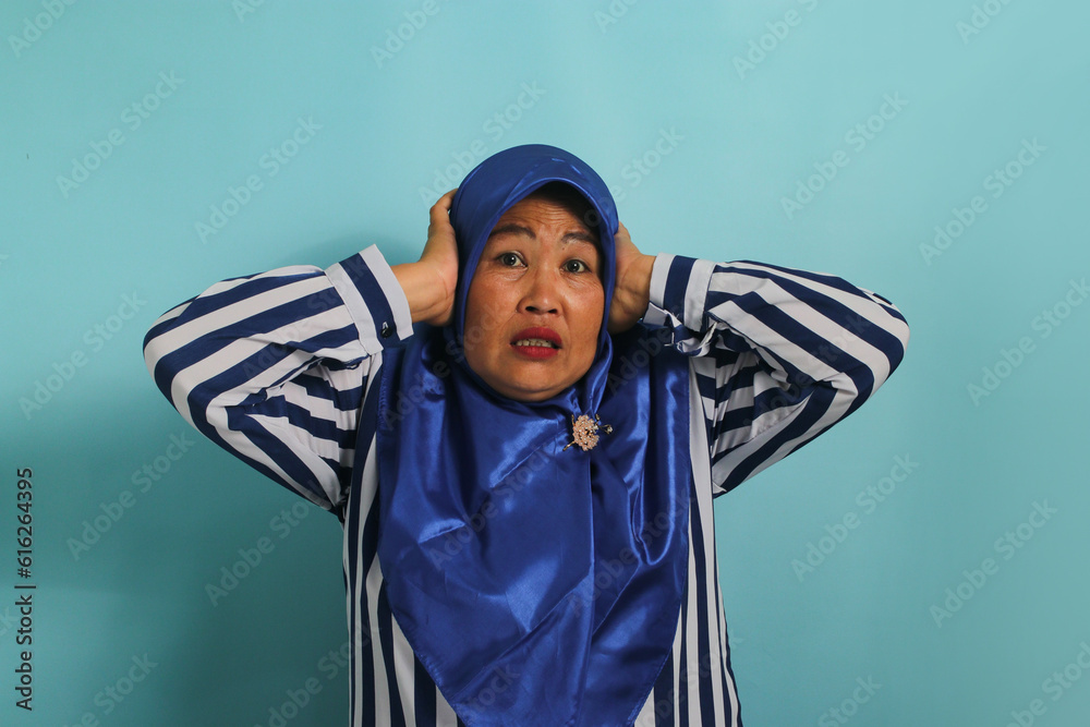 A troubled middle-aged Asian woman, wearing a blue hijab and a striped shirt, grabs her head and stares alarmed at the camera, feeling anxious while standing against a blue background A troubled middle-aged Asian woman, wearing a blue hijab and a striped shirt, grabs her head and stares alarmed at the camera, feeling anxious while standing against a blue background
