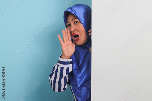 A middle-aged Asian woman in a hijab, amazed, hides behind a white wall with a blank space, peeking out and shouting next to a white advertising board for text design or advertisement.