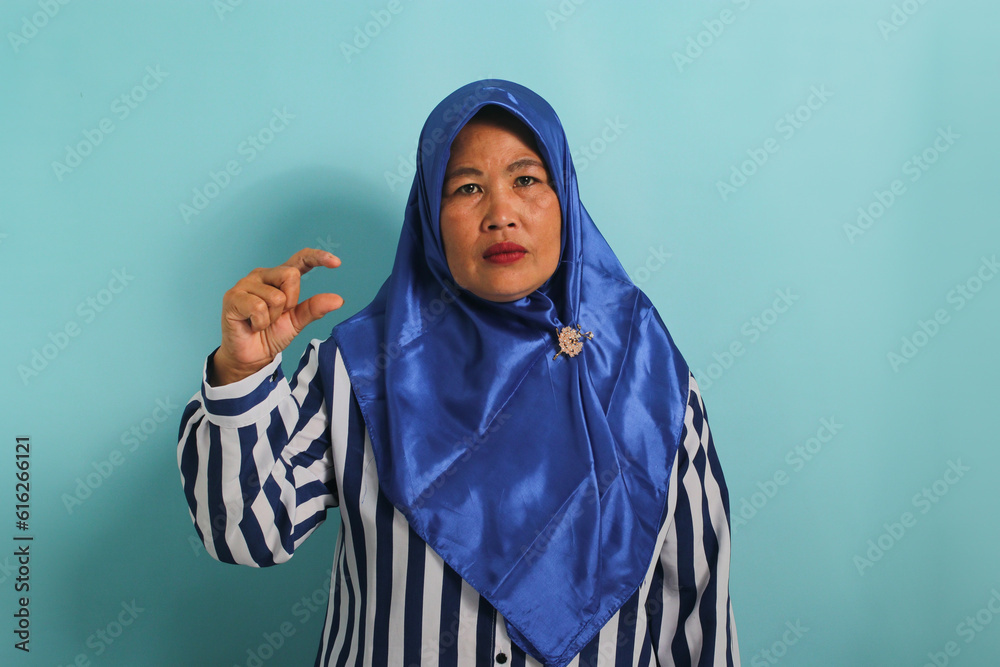 A displeased middle-aged Asian woman in a blue hijab and striped shirt shows a small gesture, looking disappointed at something very tiny in size, while standing against a blue background. A displeased middle-aged Asian woman in a blue hijab and striped shirt shows a small gesture, looking disappointed at something very tiny in size, while standing against a blue background.