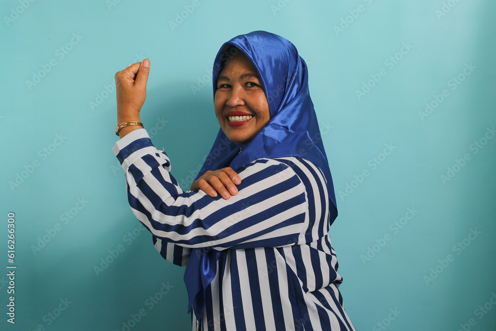 An excited middle-aged Asian woman in a blue hijab and striped shirt shows a strong gesture by lifting her arms and flexing her muscles, smiling proudly at the camera, isolated on blue background. An excited middle-aged Asian woman in a blue hijab and striped shirt shows a strong gesture by lifting her arms and flexing her muscles, smiling proudly at the camera, isolated on blue background.