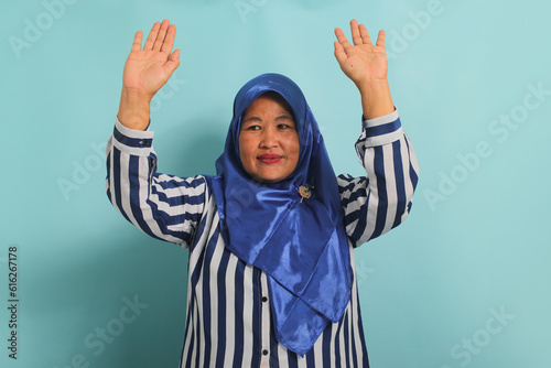 An enthusiastic middle-aged Asian woman, in a blue hijab and a striped shirt, is saying YES with a happy expression, celebrating victory with a fist pump gesture while standing over a blue background © Jamaludinyusup