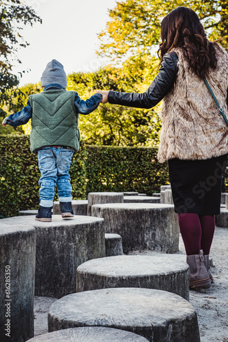 Mother helping her son move his first steps. Mom hand in hand with her boy help him learn to walk on stumps in park. Mum instills confidence to her child leading the kid through a lifelong growth path