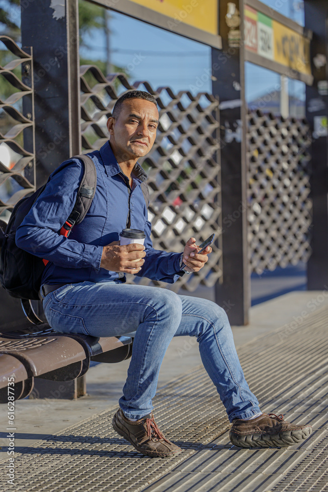 Young latin man sitting waiting for the bus with a paper cup of coffee in his hand. Young latin man sitting waiting for the bus with a paper cup of coffee in his hand.