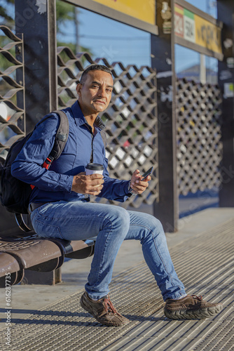Young latin man sitting waiting for the bus with a paper cup of coffee in his hand.