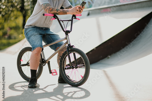 Cropped picture of a diverse tattooed man sitting on his bmx bike in a skate park and using his phone.
