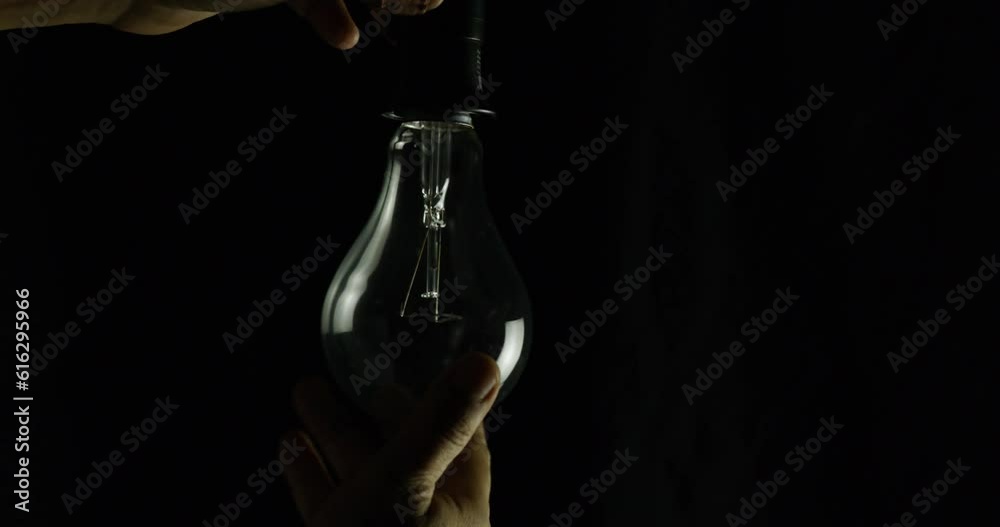 Man with his hand screws tungsten bulb into lamp base on black background.

