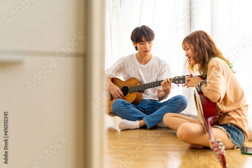 Happy young Asian man and woman friends enjoy and fun indoor leisure activity lifestyle hobbies playing guitar and singing together in living room at home on summer holiday vacation. © CandyRetriever 