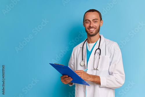 Bearded male doctor poses on blue background wearing white doctors coat, holds clipboard and pen in hands, professional people concept, copy space
