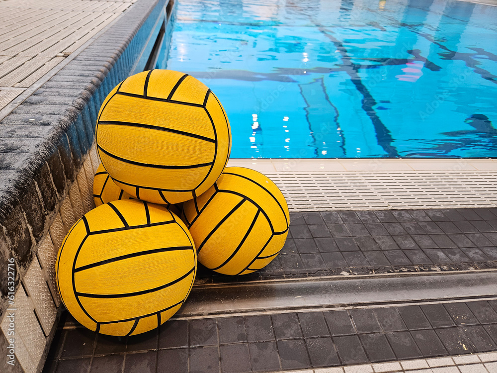 Recreation and sport image of four yellow water polo balls stacked along the edge of an indoor public swimming pool