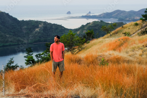 Man in Cap Enjoying the View from a Golden Grassy Hill at Sunset Overlooking a Bay.