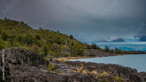 The blue glacier of Perito Moreno and turquoise Lake Argentino. In the foreground is a rocky hillside with sparse vegetation. Cloudy. Argentina. El Calafate.