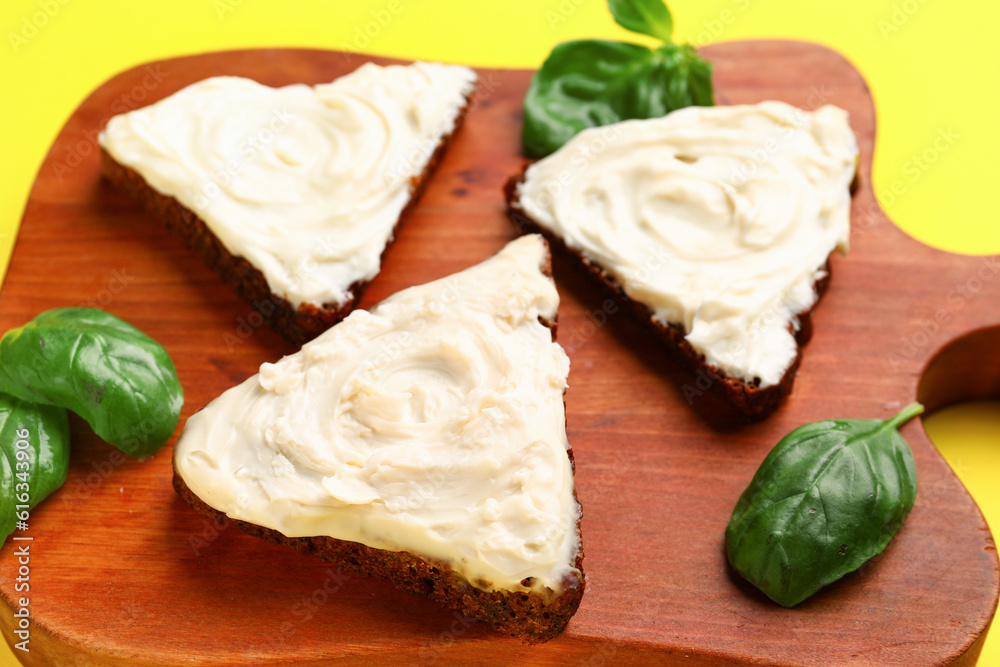 Wooden board of tasty sandwiches with cream cheese on yellow background