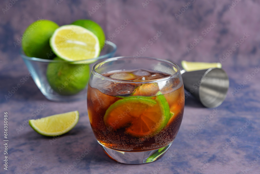 Glass of cold Cuba Libre cocktail and bowl with limes on purple background Glass of cold Cuba Libre cocktail and bowl with limes on purple background