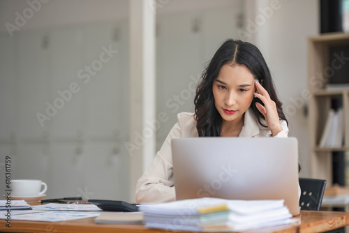 A thoughtful Asian businesswoman focuses on her tasks on her laptop, looking at her laptop with a serious face, and working at her desk in the office.