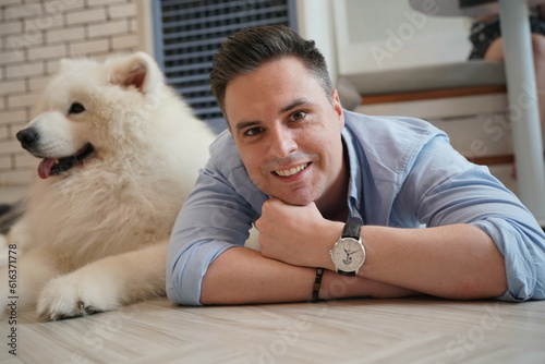 Man and Fluffy Dog Relaxing Indoors: A smiling man with a light blue shirt and a white fluffy dog lie down together on the floor.