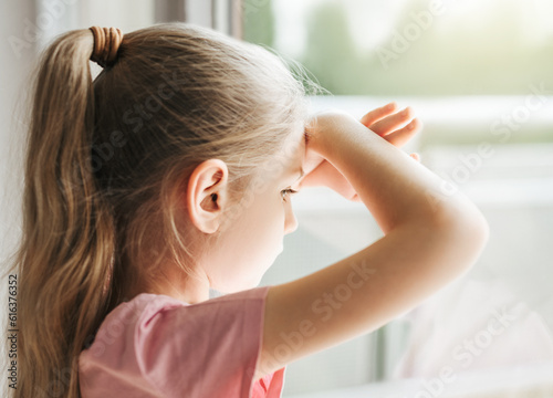 Sad little girl looking through window at home.