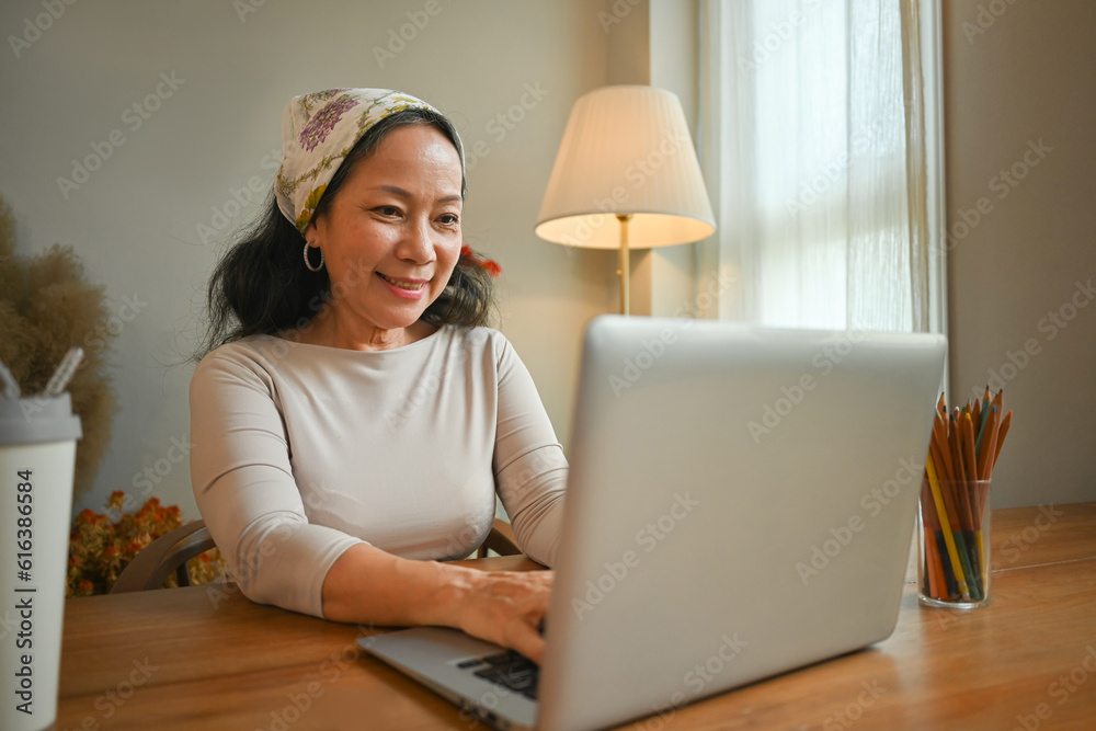 Image of happy middle age woman working online or communication on social media via laptop at home Image of happy middle age woman working online or communication on social media via laptop at home