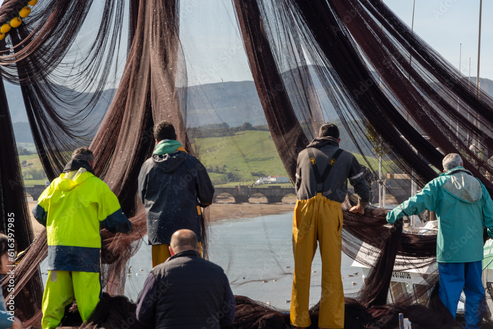 Sailors on their backs working in their work clothes repairing fishing nets in the port of San Vicente de la Barquera, a town in Cantabria, with its characteristic stone bridge in the background.