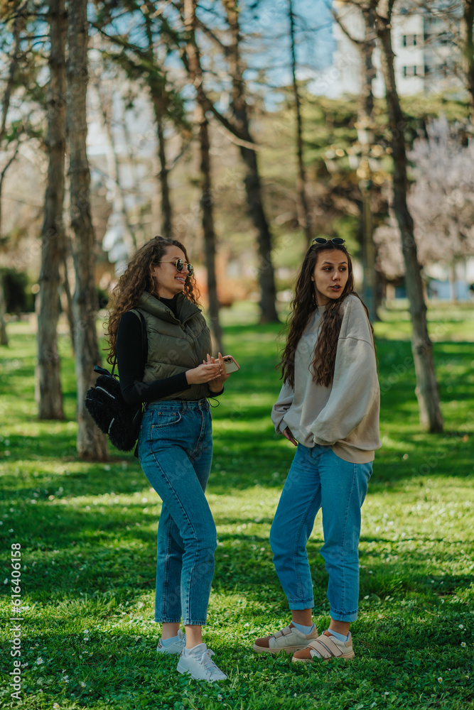 Full length photo of two lovely girls standing on a grass in the park. Brunette girl was distracted by something while talking to her friend Full length photo of two lovely girls standing on a grass in the park. Brunette girl was distracted by something while talking to her friend