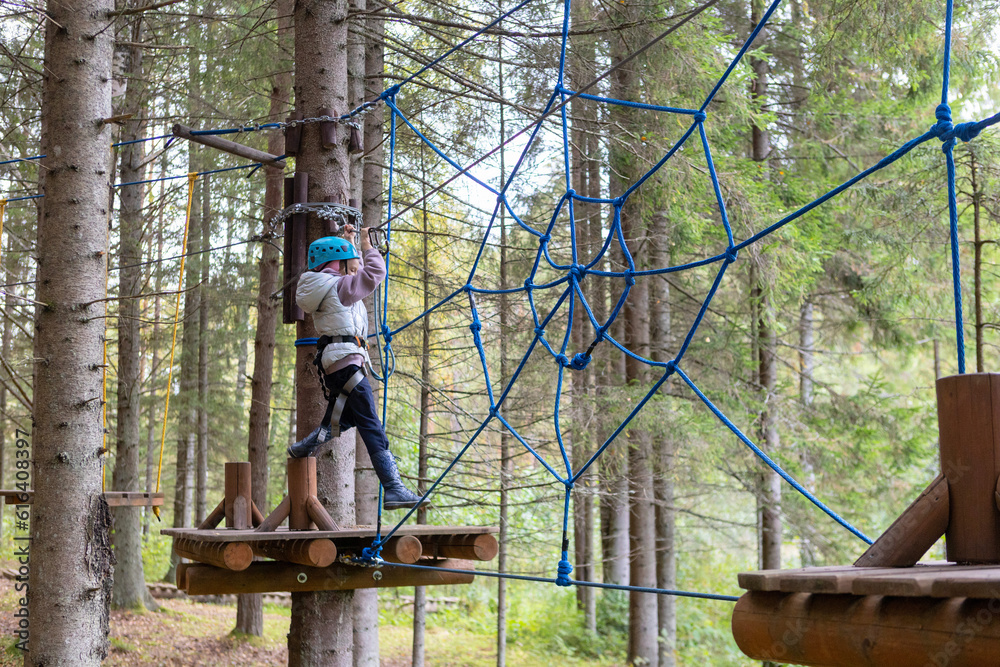 A child wearing a helmet and safety ropes will pass rope trails in a sports park.Rope town in the forest.A girl overcomes obstacles in a rope park among trees.Outdoor activities