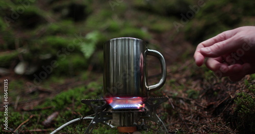 A metal mug is heated on a gas burner in the forest outdoors during a traveler's vacation. The concept of making tea in solo travels.
