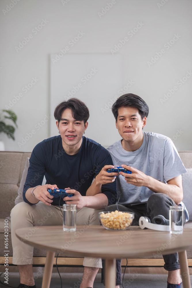 Two excited and overjoyed young Asian male friends are enjoying playing video games on a couch in the living room together. leisure, gadget entertainment, friendship Two excited and overjoyed young Asian male friends are enjoying playing video games on a couch in the living room together. leisure, gadget entertainment, friendship