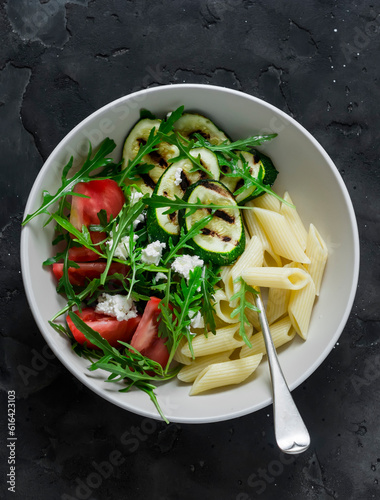 Vegetarian lunch - pasta and grilled zucchini, tomatoes, arugula, feta salad on a dark background, top view