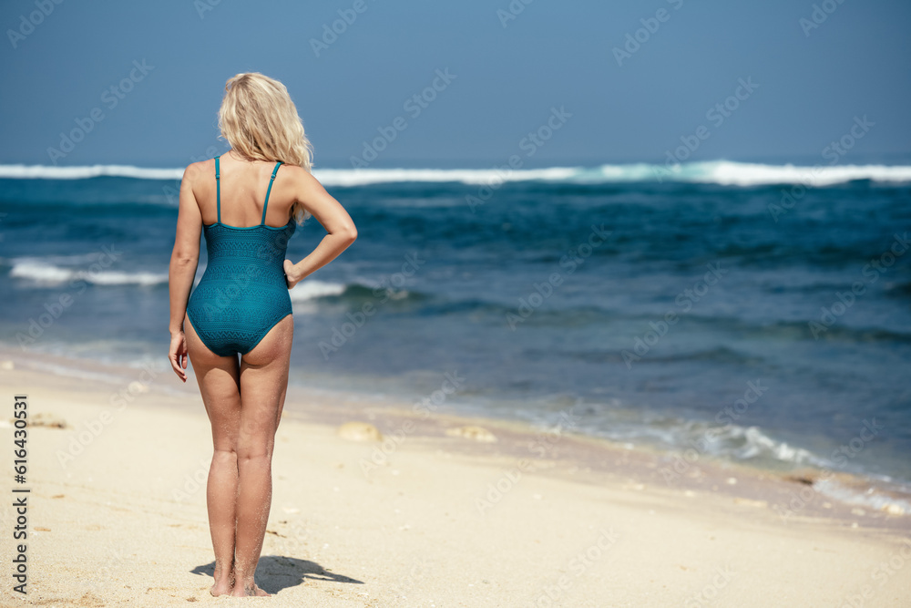 A girl in a green swimsuit stands on the beach and looks at the ocean. View of a woman's back against the background of water and sand. Beach holidays in Bali. A blonde with tanned skin enjoys.