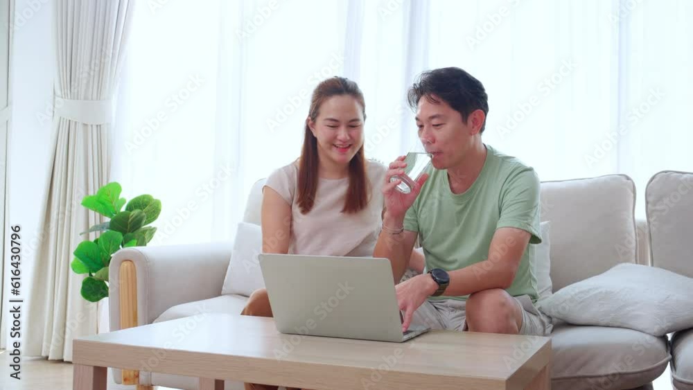 Young asian man working on laptop computer while woman giving water glass for drinking and relax sit on sofa in the living room at home, couple watching laptop online, lifestyles and family concept. Young asian man working on laptop computer while woman giving water glass for drinking and relax sit on sofa in the living room at home, couple watching laptop online, lifestyles and family concept.